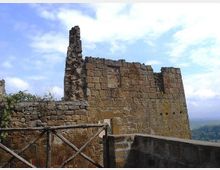 Rovine di una struttura in pietra con alti muri parzialmente crollati, affacciata su un paesaggio collinare. In primo piano si vede una terrazza con parapetto e una staccionata in legno, sotto un cielo parzialmente nuvoloso.