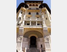 Facciata di un edificio storico a più piani vista dal basso, con ampio portale ad arco decorato a mosaico, scalinata d’ingresso e balconi sovrapposti. La struttura presenta colonne, logge ad arcate e tetto sporgente con elementi ornamentali in pietra.