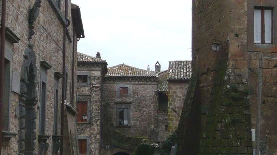 Strada acciottolata in un borgo storico, fiancheggiata da alti edifici in pietra con finestre e tetti in coppi. In fondo si vede un arco in muratura che attraversa il passaggio, con muri segnati da muschio e umidità.