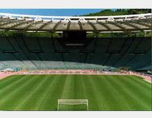 Vista dall’interno di uno stadio all’aperto con ampie tribune coperte, un grande campo in erba verde e una pista di atletica che circonda il terreno di gioco. In primo piano si vede una porta, con la copertura dello stadio e alberi sullo sfondo.