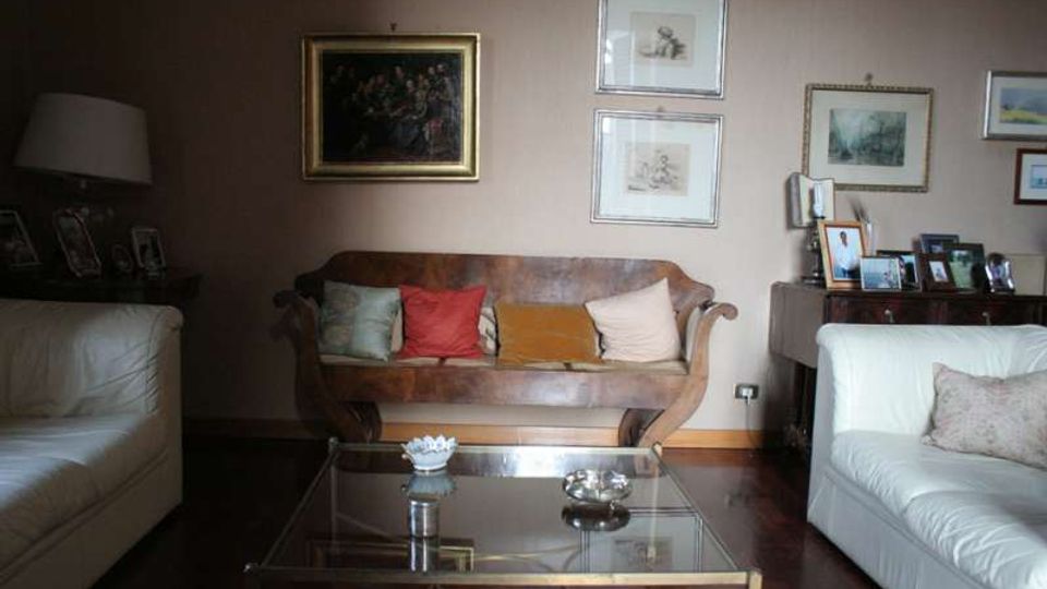 Soggiorno con divani in pelle chiara ai lati e un divano centrale in legno con cuscini colorati, davanti a un tavolino basso in vetro. Pareti beige decorate con quadri e stampe incorniciate e un mobile laterale con cornici fotografiche.