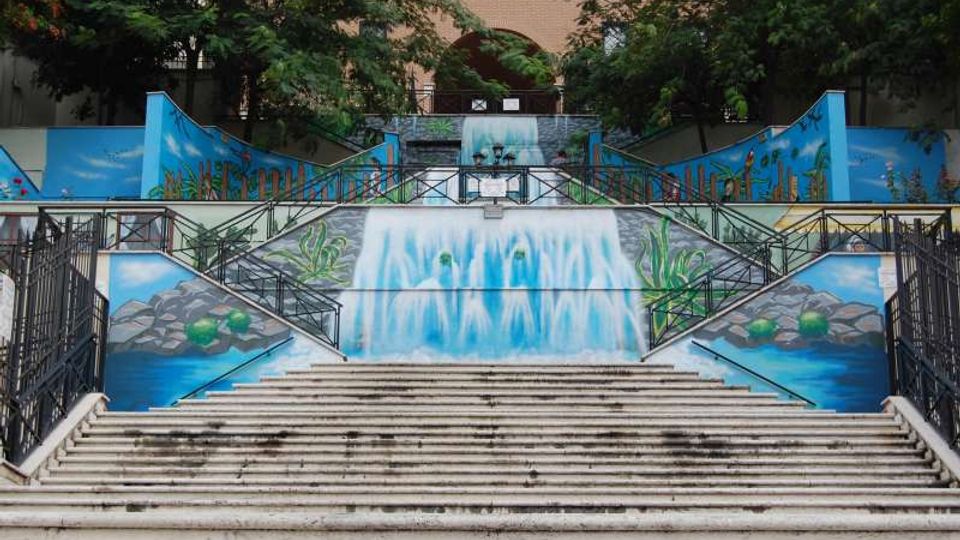 Scalinata urbana in cemento con corrimano metallici, fiancheggiata da grandi murales azzurri che raffigurano una cascata, acqua e rocce. In cima si vedono alberi e la facciata di un edificio in mattoni sullo sfondo.