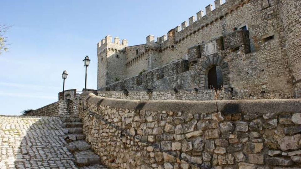 Castello medievale in pietra con mura merlate e torre, visto da una strada in salita lastricata e affiancata da un muretto in pietra e lampioni. Un grande arco d’ingresso si apre nella fortificazione sotto un cielo sereno.