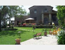 Cortile-giardino con ampio prato curato, vialetto in pietra e vasi di piante, con panchina e piccoli tavolini da esterno. Sullo sfondo si vede un edificio rurale in pietra a due piani con terrazza, scala esterna e un patio con ombrellone, circondato da alberi.
