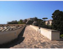 Lungo camminamento sopraelevato in pietra, affiancato da muretti chiari con copertura in coppi su un lato. Sullo sfondo si vedono edifici bassi e alberi mediterranei, tra cui palme, sotto un cielo sereno.