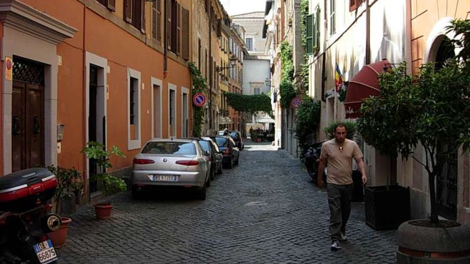 Strada stretta in pavé con auto parcheggiate ai lati, fiancheggiata da palazzi alti con facciate colorate, persiane e lampioni. Vasi e grandi fioriere con alberelli costeggiano i marciapiedi e la via prosegue in prospettiva verso un edificio sullo sfondo.