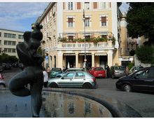Piazza urbana con una fontana circolare in primo piano e una statua scura al centro, circondata da auto parcheggiate. Sullo sfondo si vede un edificio a più piani color giallo chiaro con balcone e portico ad archi, affiancato da altri palazzi e alberi.