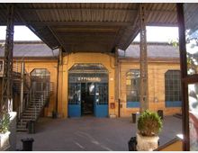 Cortile interno di un complesso industriale con tettoia in lamiera sostenuta da pilastri metallici e una grande porta vetrata blu con insegna “OFFICINE”. Ai lati si vedono finestre ad arco, una scala metallica esterna e muri in mattoni gialli.
