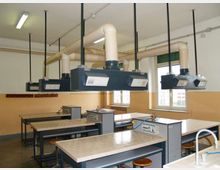 Aula laboratorio con banchi da lavoro rivestiti in piastrelle e sgabelli, dotata di cappe aspiranti sospese collegate a grandi condotti di ventilazione. Sulle pareti chiare ci sono finestre e illuminazione a soffitto, con impianti e tubazioni visibili.