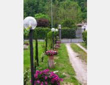 Vialetto ghiaioso in un giardino con prato, vasi di fiori e lampioni con sfere trasparenti, che conduce a un cancello metallico. Sullo sfondo si vedono alberi fitti su un pendio e alcune case con tetti in tegole.