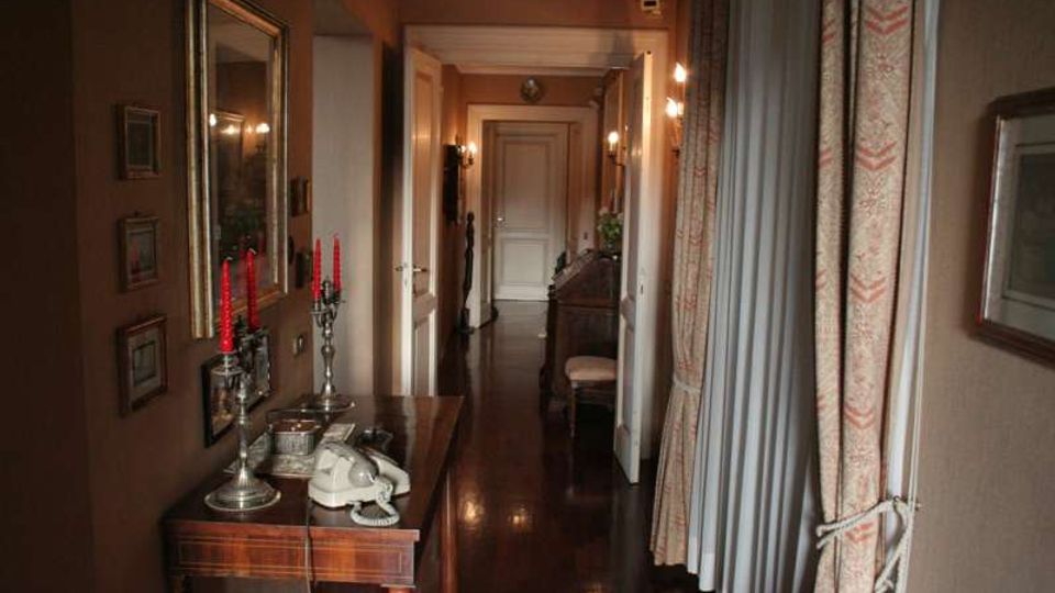 Lungo corridoio interno con pavimento in legno lucido e pareti color beige, illuminato da applique e con porte bianche lungo i lati. In primo piano un tavolino in legno con specchio, candelabri e telefono, mentre sul lato destro ci sono tende pesanti davanti alle finestre.
