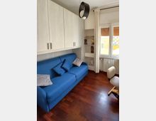 Soggiorno compatto con divano blu sotto pensili bianchi a parete, pavimento in parquet scuro e lampadario moderno. Accanto a una finestra con tende chiare e termosifone si vede una nicchia con mensole e una piccola sedia imbottita.