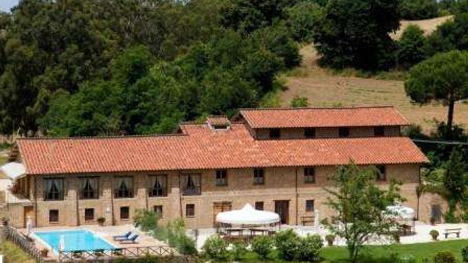 Complesso rurale in pietra con lungo edificio a due piani e tetto in tegole rosse, immerso in un paesaggio collinare con bosco sullo sfondo. In primo piano si vedono una piscina recintata, un’area esterna con ombrelloni bianchi e giardino con alberi e prato.