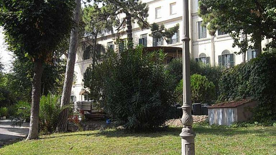 Lampione in ferro in un’area verde con prato, alberi alti e cespugli, delimitata da un muretto in pietra. Sullo sfondo si vede un edificio chiaro a più piani con finestre e balconi, parzialmente nascosto dalla vegetazione.