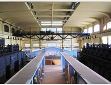 Interno di un grande edificio industriale riconvertito, con alti soffitti a volta, ampie finestre ad arco e una passerella metallica sospesa tra le pareti. Al centro si vede una lunga navata con due corridoi sopraelevati e strutture macchinari scuri ai lati.