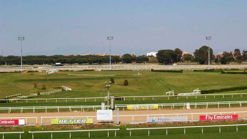 Ampio ippodromo all’aperto con pista in sabbia e piste interne erbose, delimitate da recinzioni bianche. Sullo sfondo si vedono pali per l’illuminazione, alberi e alcuni edifici lontani sotto un cielo limpido.