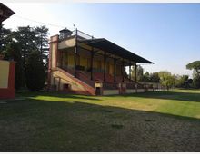 Tribuna in muratura e mattoni con gradinate e ringhiere, coperta da un’ampia tettoia metallica sostenuta da pilastri. La struttura si affaccia su un campo erboso ed è circondata da alberi in un’area verde.