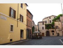 Piazzetta lastricata con edifici storici a più piani dalle facciate color ocra e rosa, con finestre a persiana e portoni ad arco. Al centro c’è un’area con tavoli e ombrelloni, e sullo sfondo si vedono edifici con balconi e vegetazione rampicante.