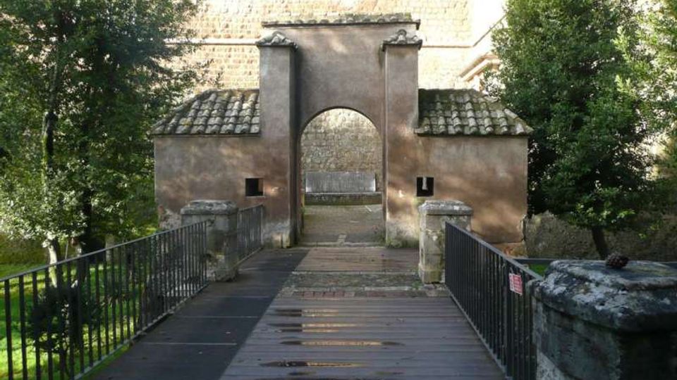 Ponte pedonale in legno con ringhiere metalliche che conduce a un ingresso ad arco in muratura, affiancato da due piccoli edifici con tetti in coppi. Sullo sfondo si vede una grande struttura in pietra, circondata da alberi e vegetazione.