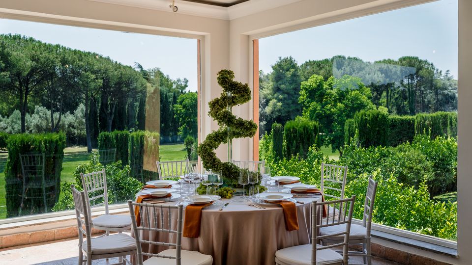 Sala da pranzo luminosa con ampie finestre panoramiche ad angolo, tavolo rotondo apparecchiato e sedie bianche. All’esterno si vede un grande giardino verde con siepi e alberi alti.
