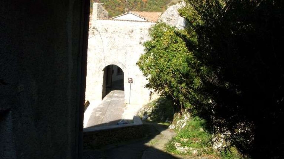 Vicolo in pendenza accanto a un edificio in pietra, con una lanterna a muro, che conduce a un arco in muratura illuminato dal sole. Sullo sfondo si vedono alberi e montagne boscose.