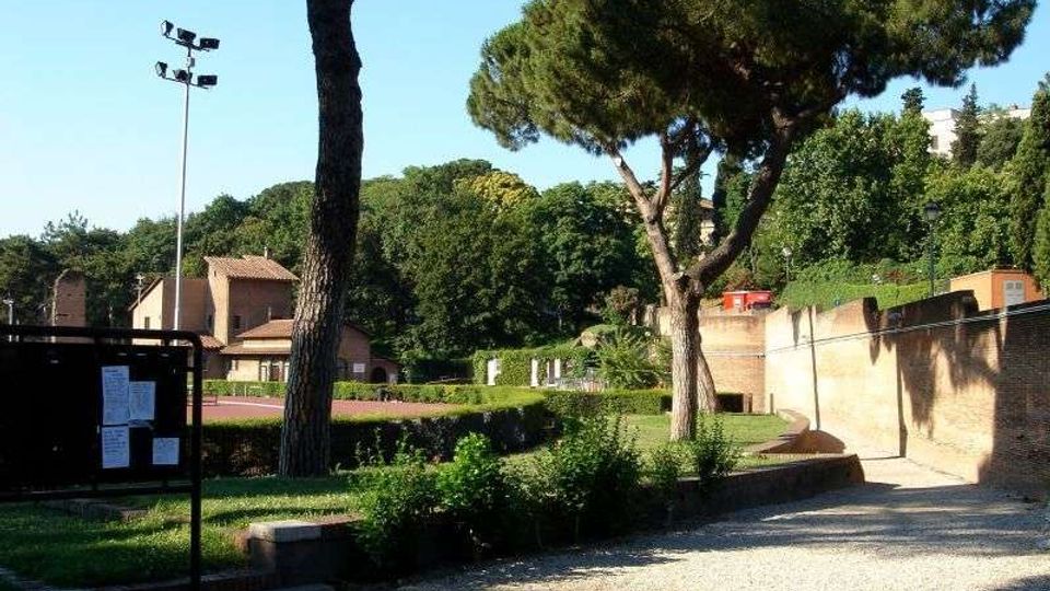 Area verde tipo parco con vialetto in ghiaia, siepi curate e grandi pini mediterranei. Sullo sfondo si vedono edifici bassi e una parete in mattoni lungo il percorso, con un palo dell’illuminazione e un pannello informativo.