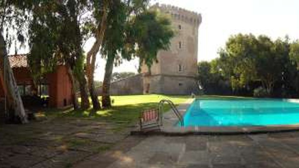Area esterna con piscina rettangolare e solarium in pietra, circondata da alberi. Sullo sfondo si vedono un edificio con tetto in tegole rosse e una torre in pietra, in un contesto verde e soleggiato.