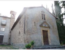 Piccola chiesa in pietra con facciata a capanna, portale in legno incorniciato da un arco e timpano decorato, e una stretta apertura verticale sopra l’ingresso. L’edificio è affiancato da un campanile in muratura e circondato da alberi alti, con un’area erbosa e vasi di piante sul davanti.