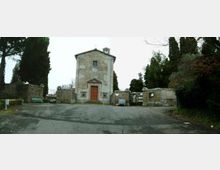 Piccola cappella in muratura intonacata con portale centrale e campanile a vela, situata all’ingresso di un cimitero con muri in pietra e cancelli laterali. Davanti si apre uno spiazzo asfaltato, circondato da cipressi e vegetazione.