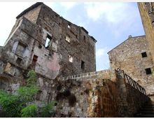 Ruderi di un edificio in pietra a più piani, con muri scrostati e finestre senza infissi, affacciati su un cortile con vegetazione. In primo piano si vede un muro di contenimento in pietra e una scala esterna con ringhiera che sale verso un altro fabbricato in muratura.