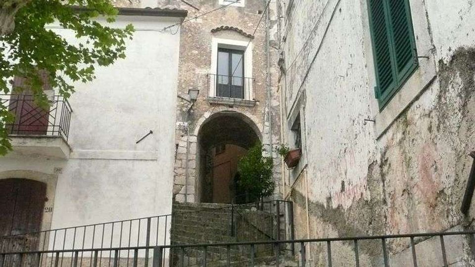 Scalinata in pietra con ringhiera metallica che sale tra edifici storici intonacati e in muratura, con arco di passaggio e balconcini. A sinistra si vede un albero e, sulla facciata a destra, una finestra con persiana verde.