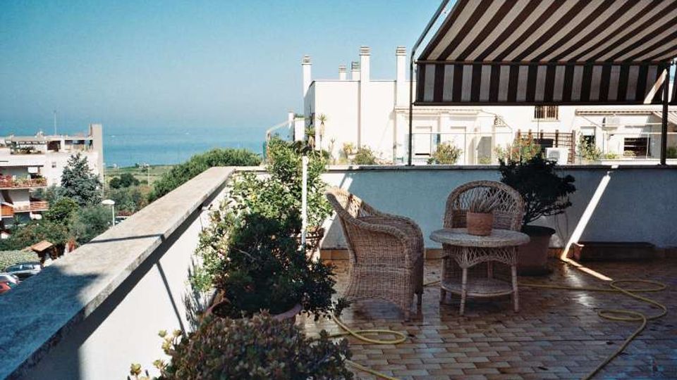 Terrazza panoramica di un edificio residenziale con pavimento in mattoni, sedie e tavolino in vimini, piante in vaso e tenda da sole a righe. Sullo sfondo si vedono altri edifici e il mare in lontananza.