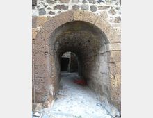 Passaggio stretto in pietra con grande arco in muratura e soffitto voltato, che conduce a un corridoio ombreggiato con pavimento in ciottoli. Le pareti sono in blocchi di pietra irregolari, con un secondo arco visibile in fondo.