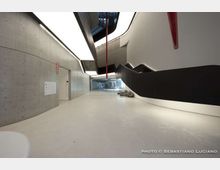 Ampio corridoio interno di un edificio moderno con pavimento chiaro, pareti in cemento a vista e illuminazione lineare a soffitto. Sul lato destro corre una grande struttura/scalinata nera e bianca dalle forme curve, con elementi rossi sospesi e uno spazio aperto visibile in fondo.