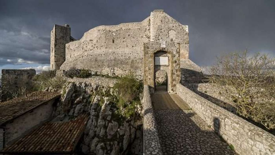 Antica fortificazione in pietra su uno sperone roccioso, con alte mura e una torre laterale sotto un cielo nuvoloso. Un ponte rialzato lastricato conduce a un portale ad arco d’ingresso, affiancato da bassi edifici con tetti in tegole.