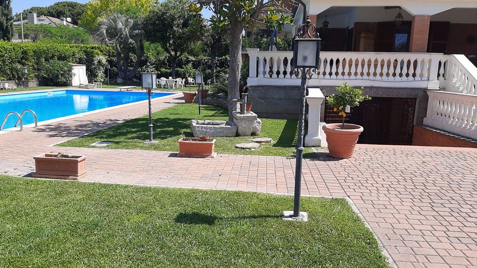Giardino di una villa con piscina rettangolare azzurra, vialetti in mattoni e prato curato con lampioni e vasi. Sullo sfondo si vede la casa con veranda coperta, ringhiera bianca e scale, circondata da alberi e siepi.