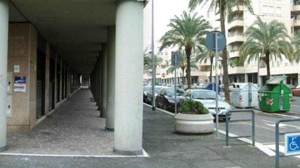 Portico urbano con lunga fila di colonne cilindriche e pavimentazione in pietra, affiancato a un marciapiede e a una strada con auto parcheggiate. Sullo sfondo si vedono palme lungo il viale, edifici residenziali alti, segnaletica stradale, un attraversamento pedonale e un parcheggio accessibile.
