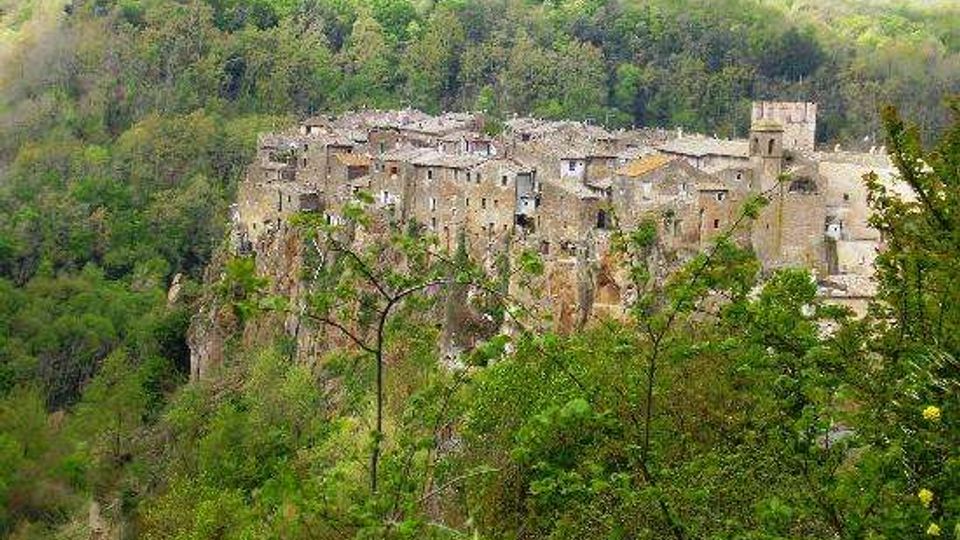 Veduta panoramica di un borgo medievale in pietra arroccato su una rupe, con case addossate tra loro e una torre quadrata. Il paese è circondato da una fitta vegetazione boschiva su pendii collinari.