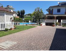 Cortile residenziale con ampia pavimentazione in autobloccanti e prato curato, con piscina rettangolare sullo sfondo. Ai lati si vedono una villetta a due piani con portico e balaustra bianca e un gazebo bianco in legno, circondati da siepi e alberi.