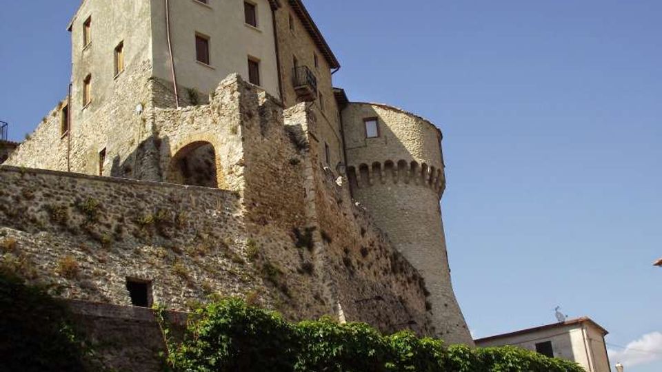Castello in pietra su uno sperone roccioso, con alte mura fortificate, archi e una torre cilindrica merlata. In primo piano vegetazione rampicante sulle pareti e cielo sereno sullo sfondo.
