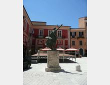 Statua in bronzo su alto piedistallo di pietra al centro di una piazza lastricata, con palco temporaneo e ombrelloni sullo sfondo. Intorno si vedono edifici a più piani con facciate colorate, balconi e finestre affacciate sulla piazza.