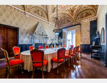 Sala da pranzo storica con grande tavolo ovale apparecchiato e sedie imbottite rosse, pavimento in legno e boiserie scura alle pareti. Soffitto a volte affrescato, grande camino in pietra scolpita, lampadario centrale e ampia finestra con tende che illumina l’ambiente.