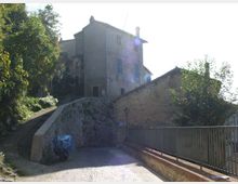 Strada in lieve salita con pavimentazione in pietra che conduce a edifici in muratura su un terrapieno, con un alto muro di contenimento. A destra c’è una ringhiera metallica lungo il bordo e vegetazione con alberi sui lati.