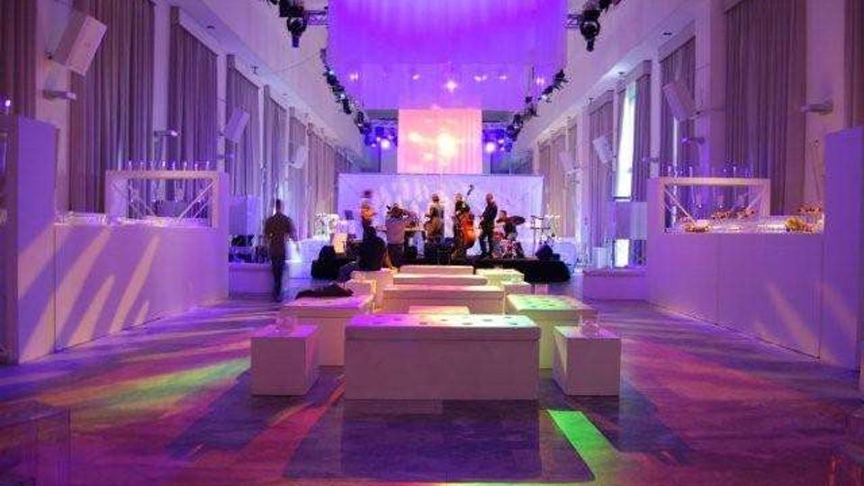 Ampia sala eventi moderna con soffitto alto, tende laterali e illuminazione scenografica viola e multicolore sul pavimento. In fondo c’è un palco con schermo e impianto luci, mentre al centro sono disposti tavoli bassi e sedute bianche e ai lati banconi espositivi.