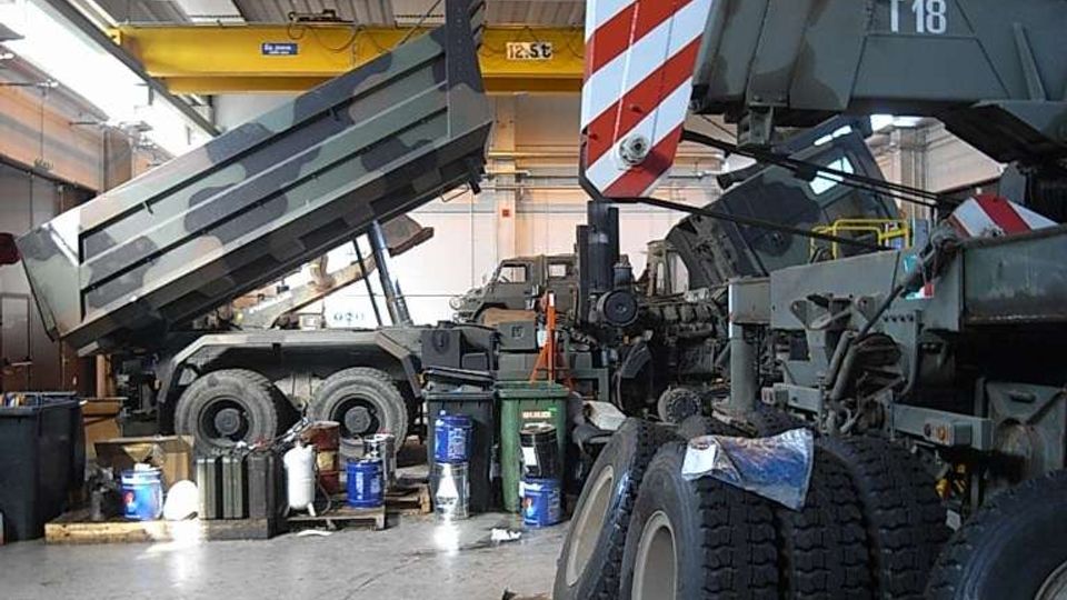 Interno di un’officina industriale con tetto metallico e illuminazione a soffitto, dove si vedono un camion ribaltabile con cassone sollevato e un grande mezzo pesante in manutenzione. Sul pavimento in cemento sono presenti pneumatici, taniche e attrezzature da lavoro sparse.