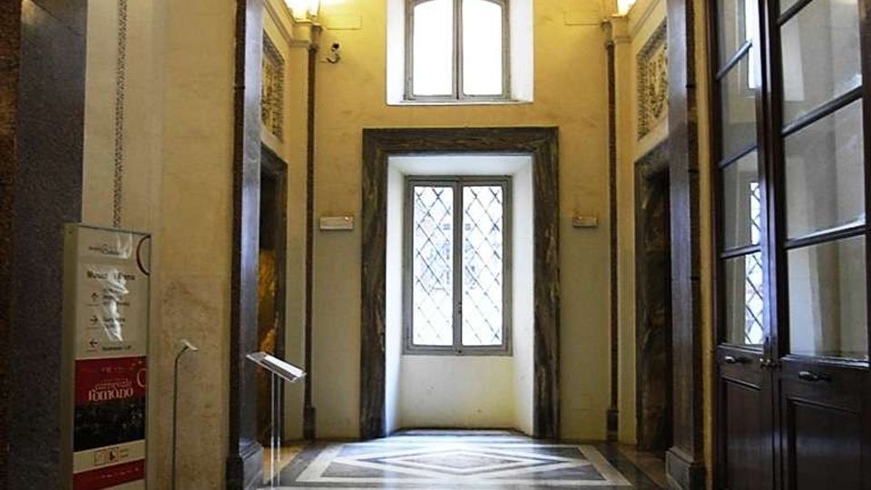 Interno di un corridoio in stile storico con soffitto a volta decorato e illuminazione a parete, pavimento in pietra con motivi geometrici e una finestra sul fondo. Sulla destra si vede una parete con grandi vetrate e porte, mentre a sinistra sono presenti colonne e un pannello informativo.