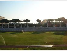 Ampio campo sportivo in erba con irrigatori in funzione e pista di atletica in primo piano. Sullo sfondo si vede una gradinata in pietra con statue allineate, alberi a chioma piatta e cielo luminoso.