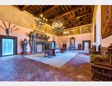 Ampia sala storica in stile rinascimentale con soffitto a travi di legno, grandi lampadari e pavimento in mattoni. Lungo le pareti ci sono un grande camino in pietra, armature decorative, affreschi e arredi d’epoca come sedie intagliate, tavoli e specchi.