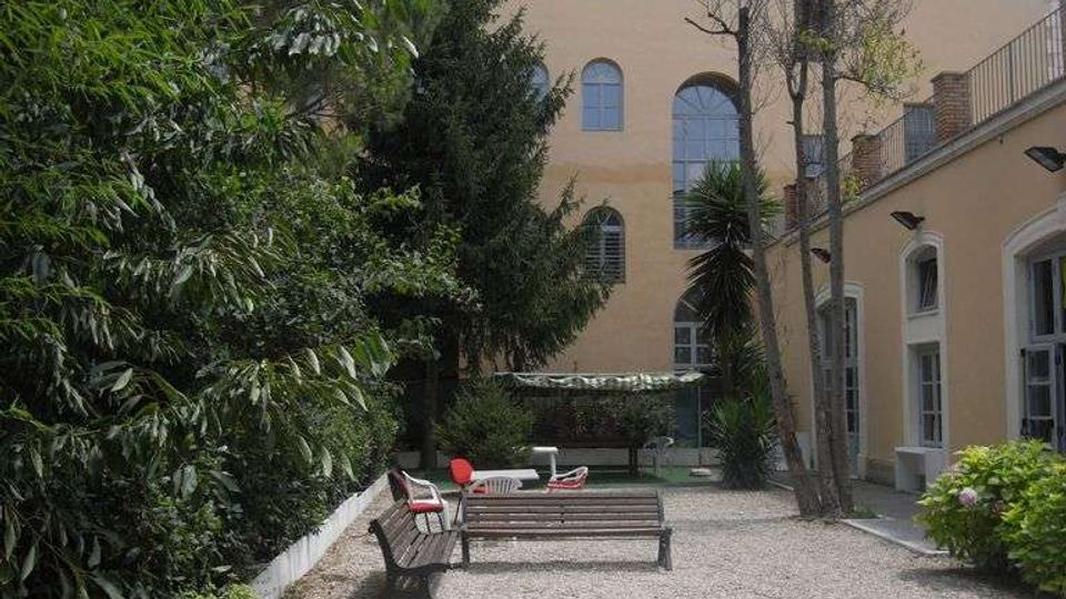 Cortile interno con pavimentazione in ghiaia, panchine e tavoli bianchi, circondato da alberi e siepi. Sullo sfondo si vede un edificio chiaro a più piani con grandi finestre ad arco e, a destra, una struttura con finestre e scala esterna.