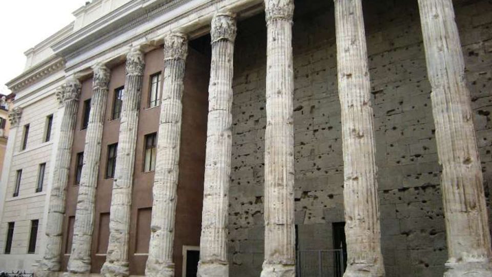 Facciata di un edificio storico con un lungo portico di colonne in pietra scanalate e capitelli decorati, allineate davanti a un grande muro in muratura. Sul lato sinistro si vede un’ala con finestre rettangolari e una recinzione metallica lungo il marciapiede.